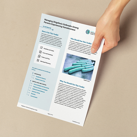 A person holds a printed page featuring an introduction to the Shigellosis Outbreak Management Toolkit, including sections on how to use the resource, who should use it, and an image of bacteria.